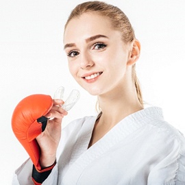 A young girl wearing a martial arts robe and boxing gloves preparing to insert a mouthguard